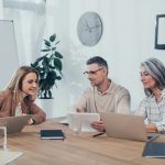 smiling multicultural colleagues talking and using gadgets in creative agency